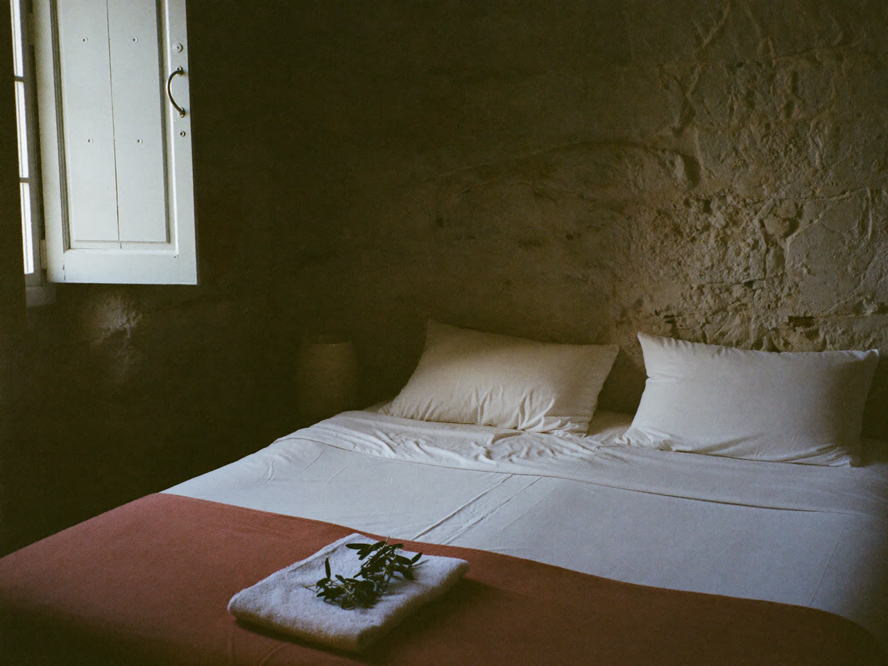 A freshly made guesthouse bed with a folded towel and olive sprig in morning light
